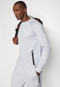 Sweatshirt técnico cinza claro com painéis laterais texturizados, fechos pretos e detalhes com logótipo no peito e na manga. Combine com calças a condizer.