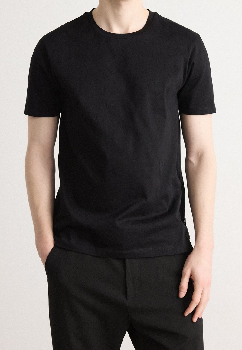 Homme portant un t-shirt noir uni à manches courtes et un pantalon noir, debout devant un fond clair avec une main dans la poche.