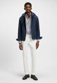 Marinblå lättviktsjacka med huva, vit skjorta, vita byxor och svarta loafers. Har synlig sömnad och uppvikt ärm.