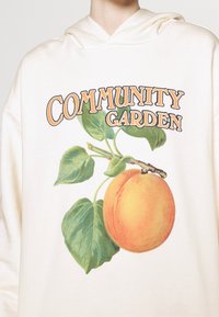 Sweat à capuche blanc avec le texte "Community Garden" au-dessus d'une illustration d'un fruit orange et de feuilles vertes sur une branche.