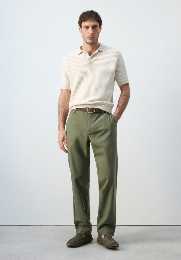 Mann steht mit einer Hand in der Tasche, trägt ein cremefarbenes kurzärmeliges Strick-Poloshirt, olivgrüne Hose, braunen Gürtel und braune Loafers.