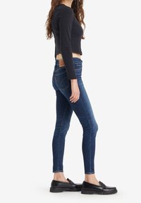 Mujer vestida con jeans ajustados de color azul oscuro, top corto de manga larga negro y mocasines negros, de perfil lateral contra un fondo blanco.