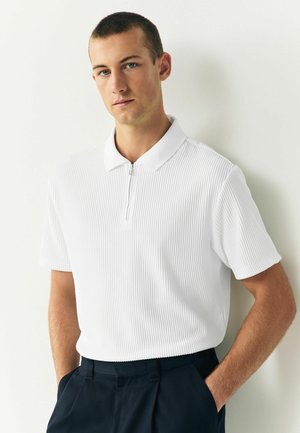 Weißes Poloshirt mit kurzen Ärmeln, gerippter Textur und einem Reißverschluss auf der Vorderseite. Kombiniert mit dunkelblauen Hosen. Einfaches Kragen Design.