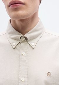 Μπεζ πουκάμισο με κουμπιά και δομημένο γιακά, με λευκά κουμπιά και ένα μικρό κεντητό λογότυπο στο στήθος. Διακριτή υφή υφάσματος ορατή.