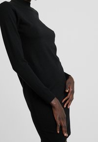 Robe noire à manches longues avec col montant, en tissu tricoté. Coupe ajustée, texture lisse, ourlet et poignets côtelés.