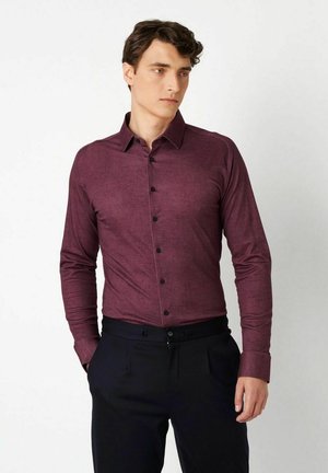 Junger Mann trägt ein eng anliegendes, kastanienbraunes Hemd mit langen Ärmeln und schwarze Hose, steht mit den Händen in den Taschen und schaut zur Seite.