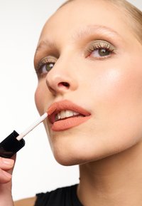 Applicatore di rossetto liquido opaco tenuto vicino alle labbra. Il prodotto presenta una morbida tonalità pesca attenuata in un elegante tubetto nero con un applicatore bianco.
