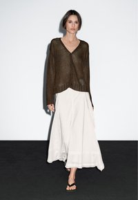 Bruine gebreide cardigan met een V-hals, knopen aan de voorkant en een geribde zoom, gecombineerd met een witte, luchtige maxi-rok en zwarte teenslippers.