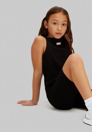 Fiatal lány hosszú barna hajjal, a padlón ülve, ujjatlan fekete Calvin Klein ruhát és fehér tornacipőt fehér zoknival viselve.