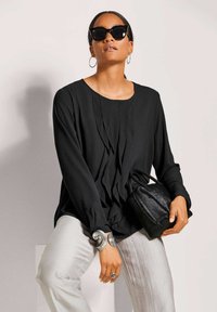 Zwarte blouse met lange mouwen en verticale plooien, gecombineerd met lichtgrijze broek. Accessoires zijn een zwarte tas en metallic sieraden.