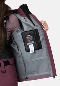 Vandtæt jakke med en dyb burgunderfarvet yderside, gråt for, netlomme med lynlås og RECCO-teknologimærke indeni.