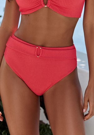Woman wearing high-waisted, textured red bikini bottoms with a matching top, standing outdoors near plants and light blue background.