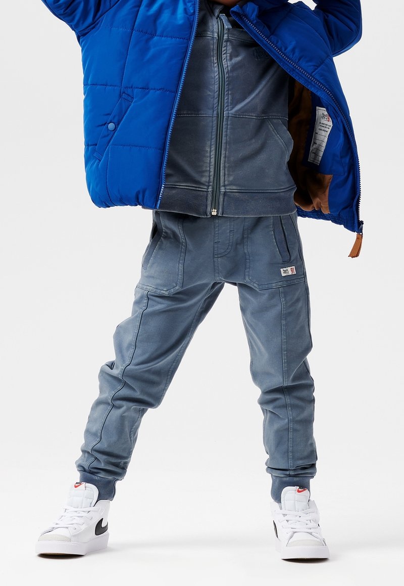 Veste matelassée bleue, pantalon style en jean gris avec poches, et baskets blanches avec des accents noirs. Coupe décontractée et confortable avec des poignets côtelés.