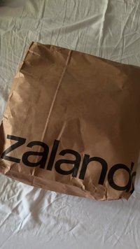 O pungă de hârtie maronie, mototolită, cu logo-ul negru "zalando" expus proeminent pe față, așezată pe o suprafață dintr-un material deschis la culoare.