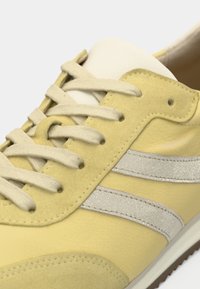 Κίτρινο αθλητικό παπούτσι με άσπρα κορδόνια, λευκή γλώσσα και δύο ανακλαστικές ασημί ρίγες κατά μήκος της πλευράς, με λεπτομερείς ραφές.
