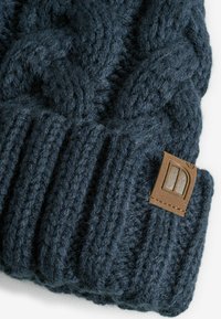 Bonnet tricoté bleu marine foncé en fil épais, arborant un motif de câble texturé et une étiquette en cuir brun avec un détail de logo.