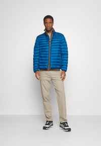 Blauwe pufferjack met horizontale stiksels, gecombineerd met een beige t-shirt met lange mouwen en lichte broek. Draag met zwarte en groene sneakers.