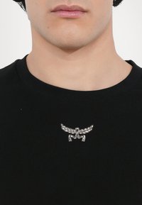 Musta T-paita, jossa on hopeinen rintaneula siipimäisellä muotoilulla ja monimutkaisella yksityiskohtaisuudella, sijoitettuna rinnan vasemmalle puolelle.