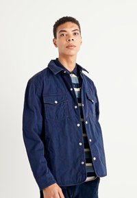 Veste matelassée navy avec couture visible, deux poches avant et fermetures à pression. Portée par-dessus une chemise rayée pour le contraste. Texture lisse.