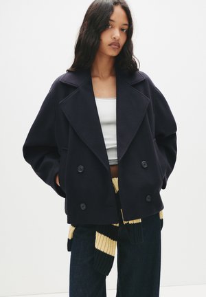 Granatowy podwójnie zapinany płaszcz marynarski z dużymi klapami, wykonany z fakturowanej tkaniny, z dwoma przednimi kieszeniami i czarnymi guzikami. Noszony na białej bluzce.