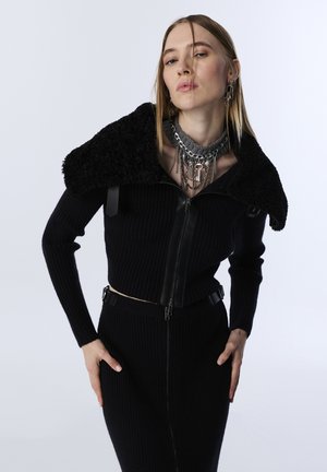 Black ribbed zip-up top with a textured faux fur collar and leather accents, paired with a matching fitted skirt, showcasing silver chain jewelry.