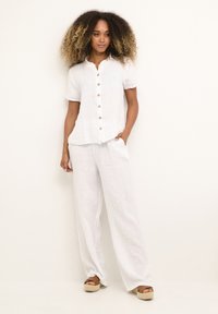 Camicia bianca di lino con maniche corte, abbinata a pantaloni larghi bianchi di lino. Le scarpe presentano una suola espadrille a strati.