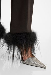 Zilveren spitse hoge hakken met steenversieringen; zwarte veerafwerking bij de enkel, gecombineerd met strakke, donkere broeken.