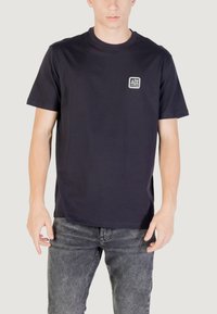 Maglietta in cotone blu navy con collo rotondo, maniche corte e una toppa con logo sul petto sinistro. Presenta un'apertura comoda.