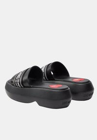 Infradito neri con piattaforma e ampie cinghie che presentano un marchio bianco. Suole testurizzate e accenti di cuori rossi sulla plantare.