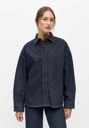 Femme portant une veste en jean foncé oversize et un pantalon assorti, debout devant un fond blanc uni.