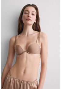 Młoda kobieta z długimi brązowymi włosami, ubrana w cielisty usztywniany biustonosz i dopasowane jedwabiste szorty, pozująca na tle białej ściany.