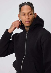 Homme aux cheveux tressés ajustant la capuche d'un sweat à capuche noir zippé, regardant sur le côté contre un fond clair uni.