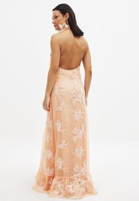Pfirsichfarbene Maxikleid mit gestickten Blumenmustern, offenem Rücken und gerafftem Saum. Aus einem leichten, fließenden Stoff gefertigt.