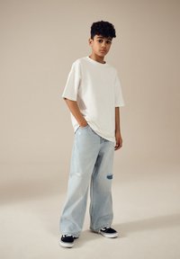 Hvid oversized t-shirt, lyseblå slidte jeans med lapper, sorte sneakers, stående mod en neutral baggrund.