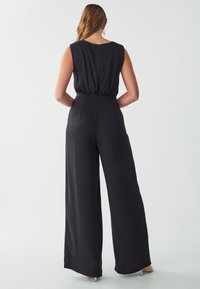 Combinaison noire sans manches avec une taille cintrée, un design à jambes larges, un tissu lisse et une fermeture éclair au dos pour un enfilage facile.