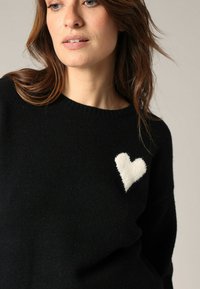 Pull noir avec une appliqué en forme de cœur blanc, doux, sur la poitrine gauche, fabriqué en matière tricotée lisse avec un col rond.