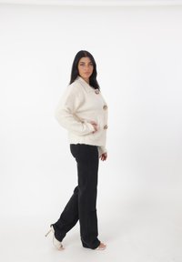 Suéter de lana texturizado en color crema con un gran cuello y tres botones decorativos, combinado con unos pantalones vaqueros negros de pierna recta y sandalias de tacón.