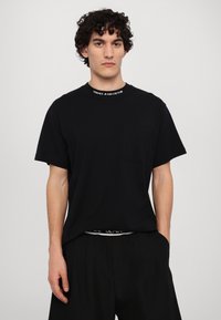 Melna T-kreklā ar apaļu kakla izgriezumu, kam ir Versace Jeans Couture zīmols. Īsas piedurknes un krūšu kabata. Mīksta auduma tekstūra.