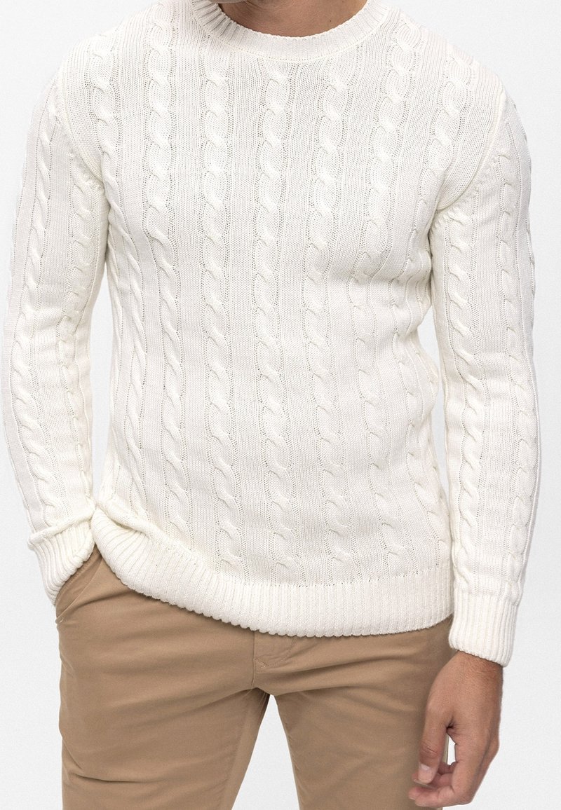 Homme portant un pull blanc en maille torsadée à manches longues et un pantalon beige, les mains partiellement dans les poches, sur un fond uni.