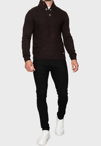 Maglione lavorato a maglia marrone con un alto colletto, due bottoni e un motivo a spina di pesce testurizzato, abbinato a pantaloni neri e sneaker bianche.