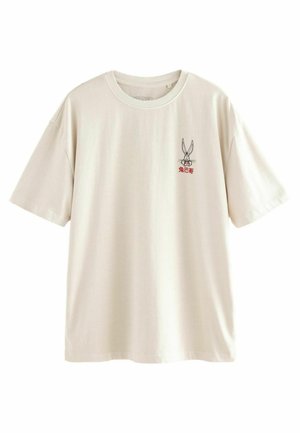 Krém színű pamut póló, rövid ujjakkal és kerek nyakkivágással. Kiemelkedő grafika egy mesebeli nyúl képével a mellkason.