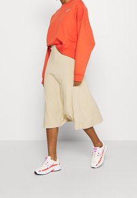 Oranges Cropped-Sweatshirt mit rundem Ausschnitt und weißem Logo, kombiniert mit einem beigefarbenen Midirock und weißen Sneakers mit rosa Akzenten.