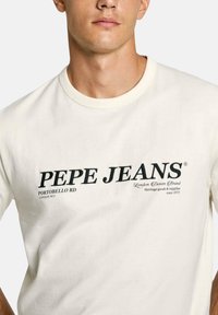 Kreem lühikeste varrukatega T-särk, millel on must "PEPE JEANS" logo ja lisatekst all. Valmistatud puuvillast, ümmarguse kaelusega.