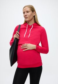 Raskaana oleva nainen, jolla on päällään punainen huppari ja mustat legginsit, kantaa mustaa olkalaukkua ja lepää kädellä vatsallaan.