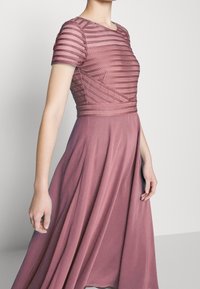 Kurzärmliges, bodenlanges Kleid in staubigem Rosa mit einem strukturierten, texturierten Oberteil und einem fließenden Rock. Mit horizontalen Streifenakzenten.
