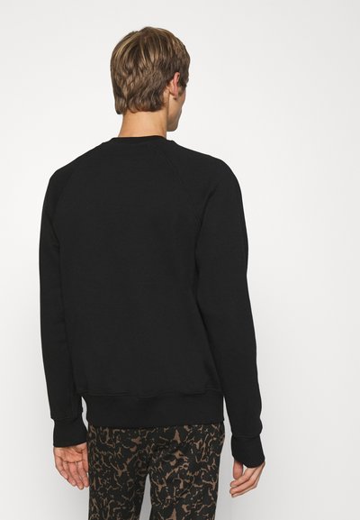 Sweatshirt noir avec des manches raglan et un col côtelé. Le matériau semble être doux et texturé. Porté par-dessus un pantalon foncé à motifs.