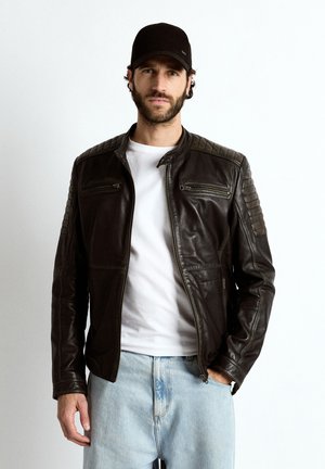 Mężczyzna w czarnej skórzanej kurtce, białej koszuli, jasnoniebieskich dżinsach i czarnej czapce stojący na białym tle.