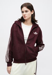 Jovem mulher vestindo uma jaqueta Adidas com fecho éclair cor vinho e riscas brancas nas mangas, por cima de uma camisa branca e calças de tom claro.