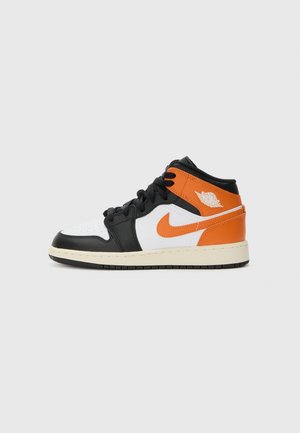 Basket montante noire, blanche et orange avec le logo Nike swoosh et le logo ailes Air Jordan sur le côté, sur un fond uni gris clair.