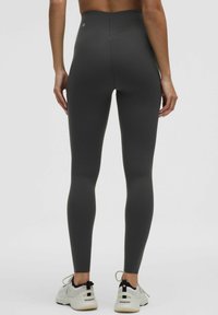 Leggings a vita alta, elasticizzati, di colore grigio scuro con una texture liscia e un logo sulla parte posteriore della vita, abbinati a sneaker chiare.